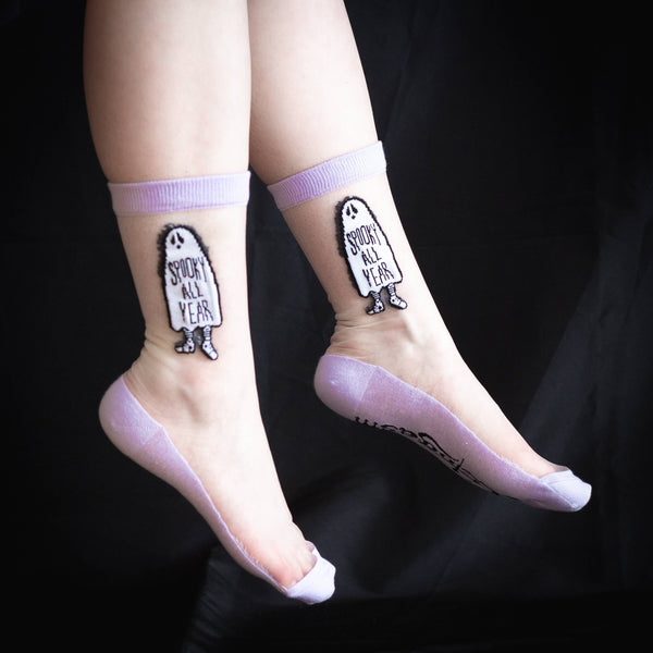 A pair of crew socks with a pale purple cuff, toe, and heel with the image of a ghost with the words “SPOOKY ALL YEAR” written on the middle of the ghost’s sheet. The brand name Ectogasm is written on the bottom of the sock , shown worn by model
