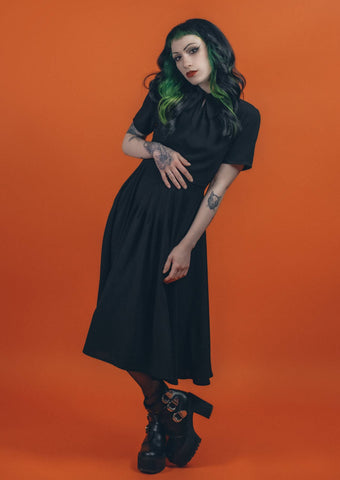 A model wearing a black 40s style short sleeved midi dress. elongated platter style collar and a narrow keyhole opening at the sternum with gathered detail and hook and eye closure. There is gathered pleating at the front of the full skirt