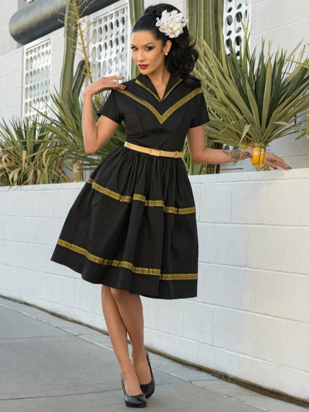 50s style Patio Dress in black stretch cotton sateen embellished with metallic gold ribbon trim features a collared v-neckline, fitted princess seamed bodice, short dolman sleeves, and gathered full  just-below-the-knee length skirt. shown on model