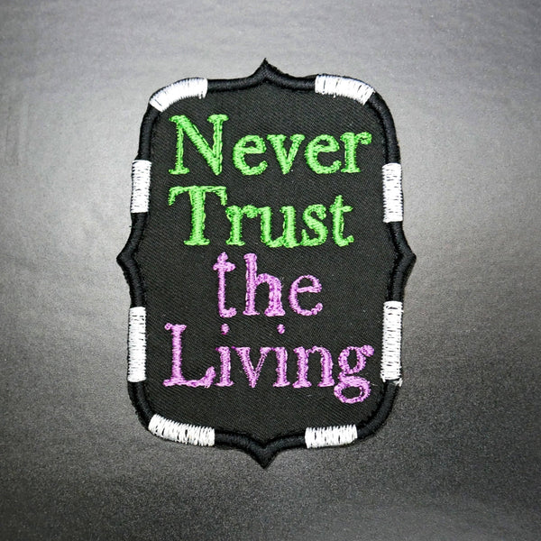 black canvas patch with "Never Trust the Living" text in green and purple with black and white stripe border
