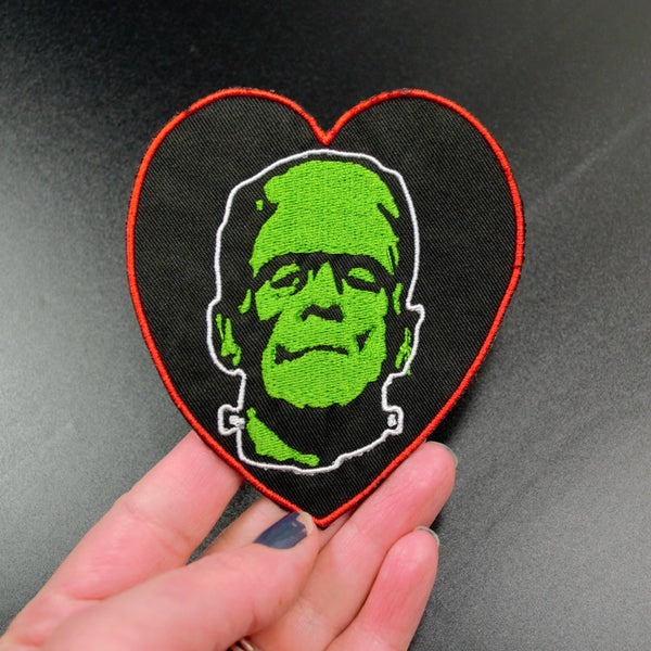 Frankenstein's monster face in green on a red bordered heart-shaped black canvas embroidered patch, held in a hand