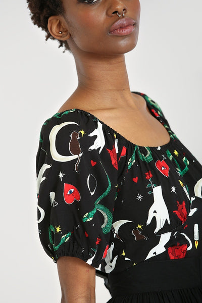 A model wearing a short sleeved peasant top in black with an all over print of snakes, crescent moons, candles, hearts, stars, and hands.