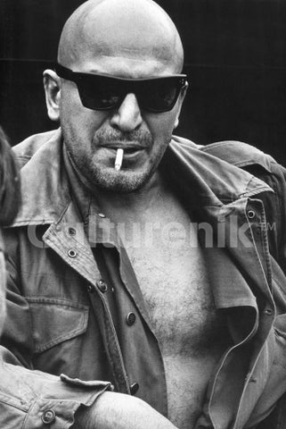 postcard of black and white photo image of Telly Savalas wearing black sunglasses with shirt open and smoking a cigarette