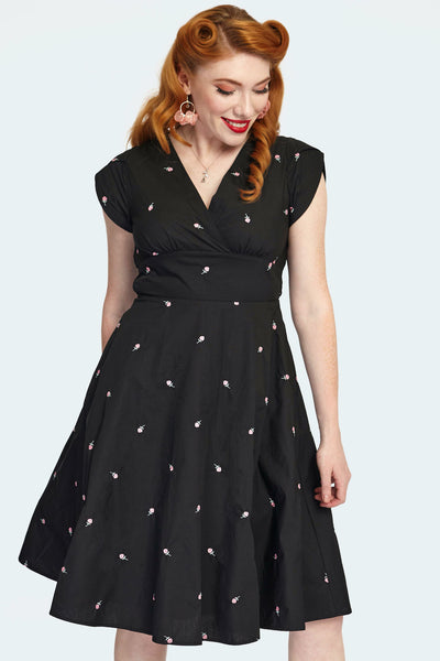 A model wearing a knee-length black fit and flare style dress with a surplice v-neckline and tulip style cap sleeves. There are small pink and green roses embroidered on the fabric of the dress. Seen in close up