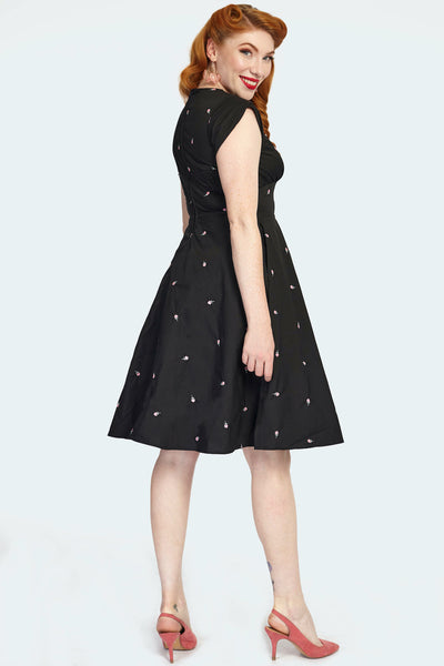 A model wearing a knee-length black fit and flare style dress with a surplice v-neckline and tulip style cap sleeves. There are small pink and green roses embroidered on the fabric of the dress. Shown from behind