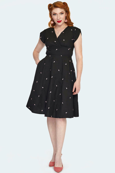 A model wearing a knee-length black fit and flare style dress with a surplice v-neckline and tulip style cap sleeves. There are small pink and green roses embroidered on the fabric of the dress. The model has one hand in a pocket of the dress