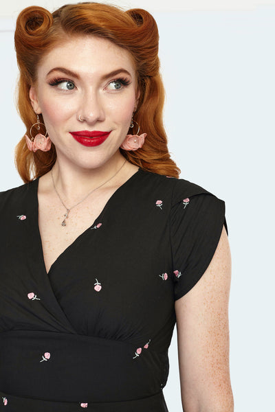 A model wearing a knee-length black fit and flare style dress with a surplice v-neckline and tulip style cap sleeves. There are small pink and green roses embroidered on the fabric of the dress. Shown in close up to feature the rose embroidery and surplice neckline