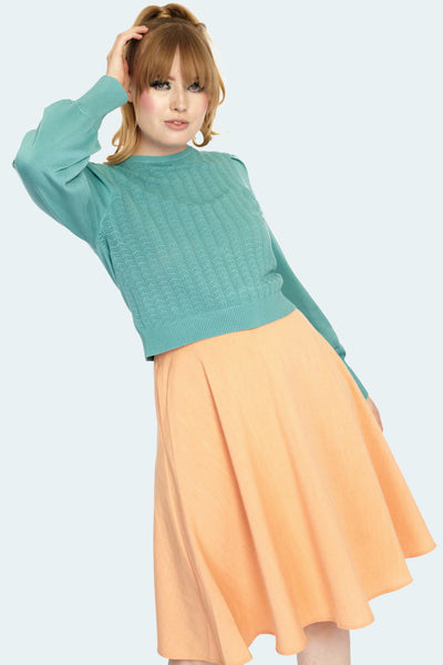 A model wearing a long sleeved pullover sweater in a robin’s egg blue color. It has a pointelle chevron style geometric pattern knitted into the front of the front of the sweater, slightly puffed shoulders with pleated detail and a ribbed jewel style neckline with matching wide ribbed cuffs. Model is shown with one arm raised