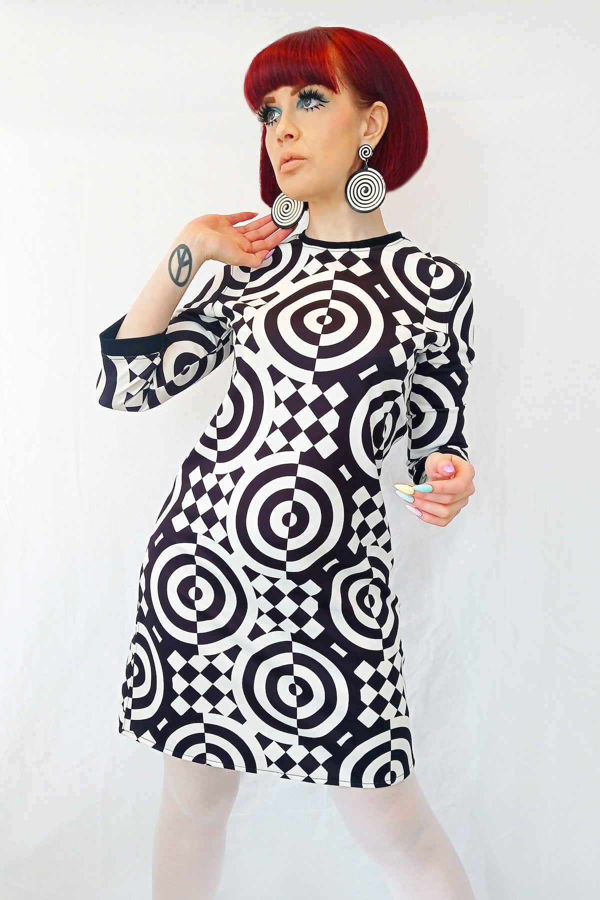A model wearing a 3/4 sleeve mini dress with a black and white checkered and concentric circle op-art style pattern. It has a round neckline