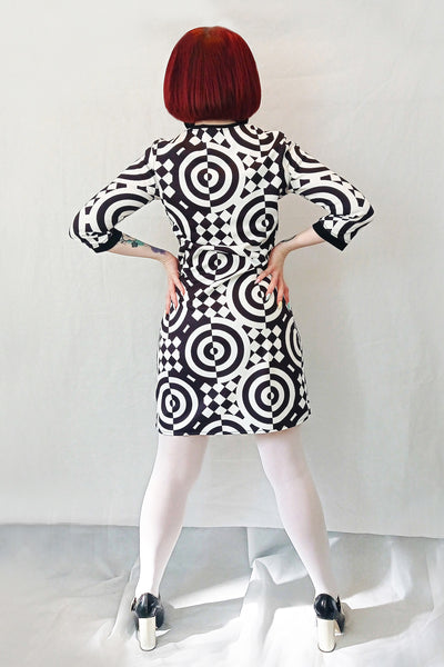 The back view of a model wearing a 3/4 sleeve mini dress with a black and white checkered and concentric circle op-art style pattern. It has a round neckline