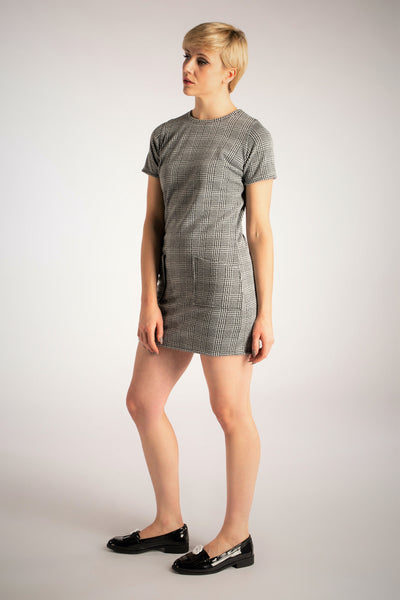 A full length shot of model wearing a short sleeved micro-mini jersey dress in a grey tartan style. It has two pockets. The shot better shows the length of the dress (high thigh length)
