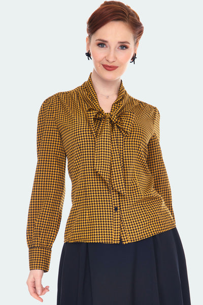 A model wearing a button up long sleeved blouse with a tie-front neck in a mustard and black houndstooth pattern. It has slightly puffed shoulders and subtle balloon sleeves