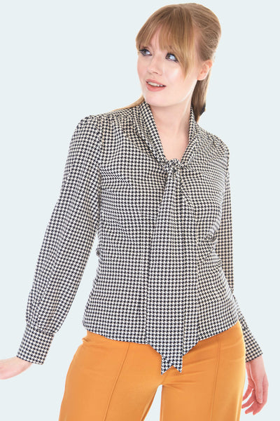 A model wearing a tie-neck blouse in a white and black houndstooth pattern. It has slightly puffed shoulders and subtle balloon sleeves.
