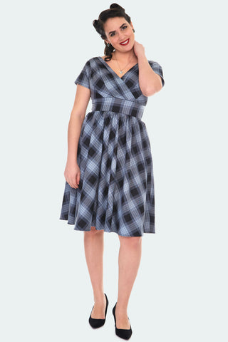 A model wearing a knee length fit and flare dress in grey and black shadow plaid. It has short sleeves, a v neck, and gathering at the bust
