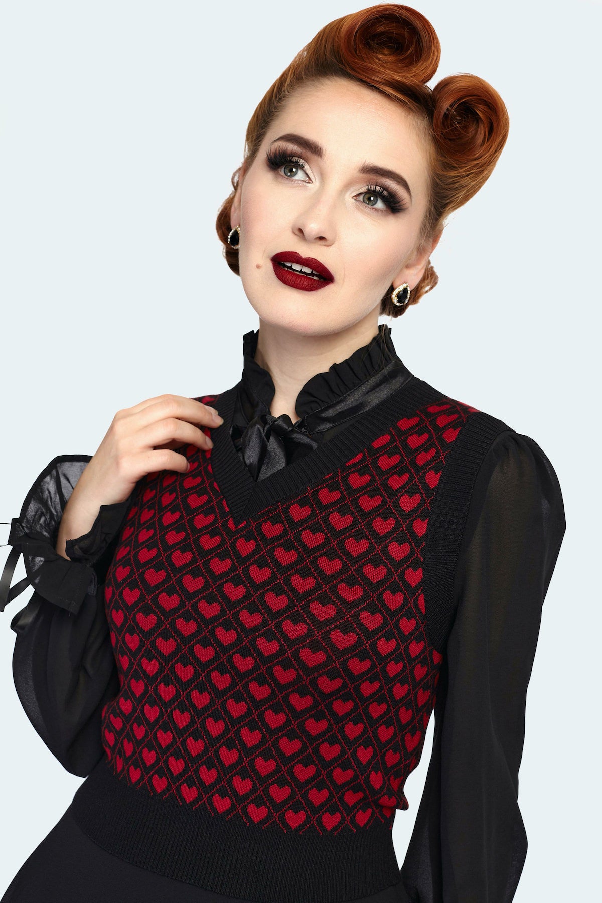 A model wearing a slightly cropped black sweater vest with a red heart and lattice pattern. It has a black ribbed v-neckline and ribbed bottom and armhole hems