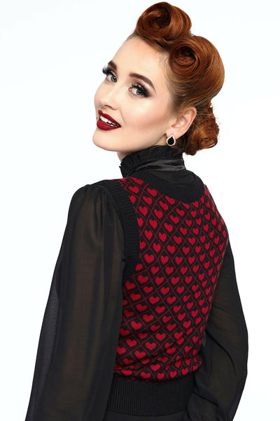 A model wearing a slightly cropped black sweater vest with a red heart and lattice pattern. It has a black ribbed v-neckline and ribbed bottom and armhole hems. Shown at a 3/4 angle from behind to show the all-over pattern