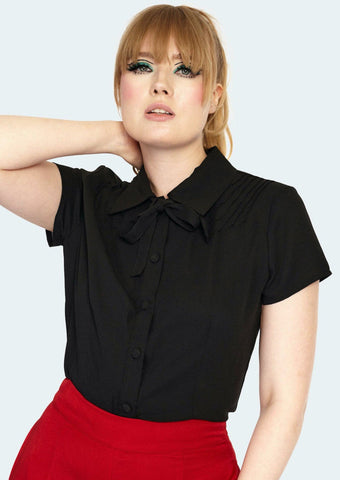 A model wearing a black short sleeved blouse with pin tuck detail on either side of the front shoulder of the blouse. It has matching black fabric buttons and a tie at the collar. Model wears blouse tucked into pants