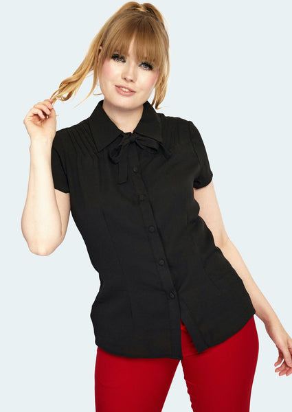 A model wearing a black short sleeved blouse with pin tuck detail on either side of the front shoulder of the blouse. It has matching black fabric buttons and a tie at the collar. Model wears the blouse untucked with pants 