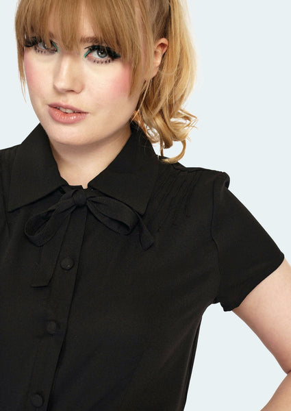 A model wearing a black short sleeved blouse with pin tuck detail on either side of the front shoulder of the blouse. It has matching black fabric buttons and a tie at the collar. Shown in close-up to better illustrate pin tuck and collar tie detail.