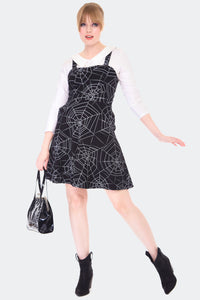A model wearing a black overall style flared dress with a white all over spiderweb pattern. She is wearing a white collared top underneath 