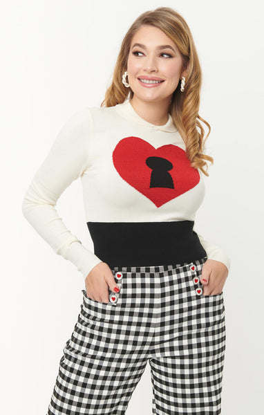 A model wearing an ivory colored intarsia knit pullover sweater with a large red heart in the middle with a black keyhole. It has an ivory Peter Pan collar, ribbed cuffs, and a wide black bottom band. 