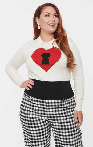 A plus size model wearing an ivory colored intarsia knit pullover sweater with a large red heart in the middle with a black keyhole. It has an ivory Peter Pan collar, ribbed cuffs, and a wide black bottom band