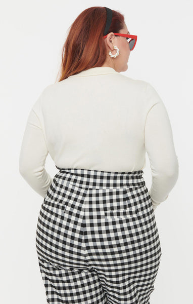 A plus size model wearing an ivory colored intarsia knit pullover sweater with a large red heart in the middle with a black keyhole. It has an ivory Peter Pan collar, ribbed cuffs, and a wide black bottom band. Shown from behind tucked into pants 