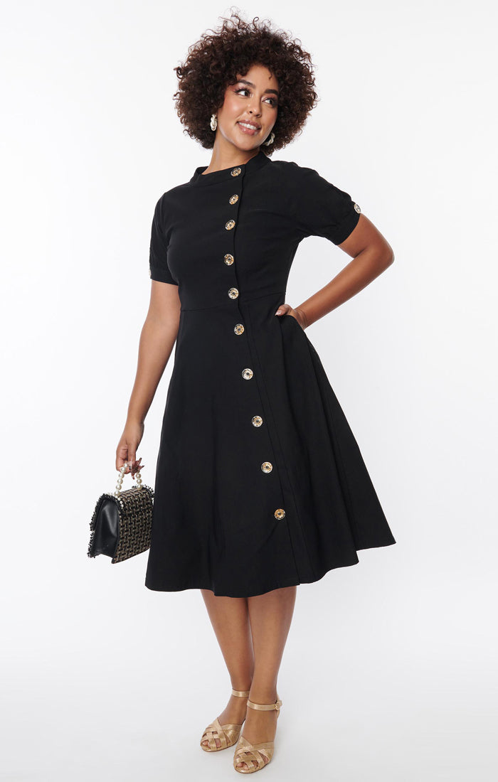 A black short sleeved dress with a high neck and full skirt. It has clear buttons with gold starburst detail down the front and on each sleeve. Shown on a model 