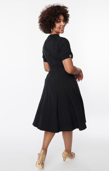 A black short sleeved dress with a high neck and full skirt. It has clear buttons with gold starburst detail down the front and on each sleeve. Shown on a model from behind 