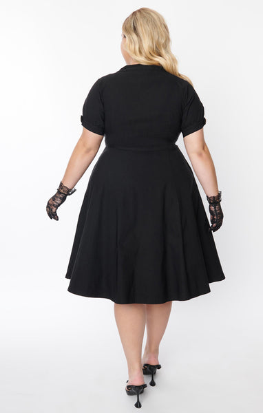 A black short sleeved dress with a high neck and full skirt. It has clear buttons with gold starburst detail down the front and on each sleeve. Shown on a plus sized model from behind