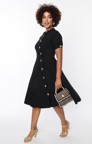 A black short sleeved dress with a high neck and full skirt. It has clear buttons with gold starburst detail down the front and on each sleeve. Shown on a model in a 3/4 shot